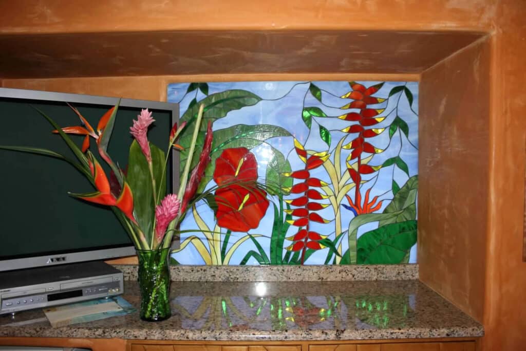 A rectangular stained glass panel featuring vibrant red anthurium flowers, red and yellow heliconia, and lush green tropical foliage against a light blue sky.