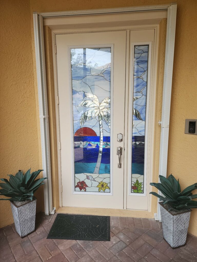 A cream-colored double entry door featuring leaded stained glass panels depicting a tropical sunset scene with a palm tree, red sun, blue ocean waves, and colorful flowers.