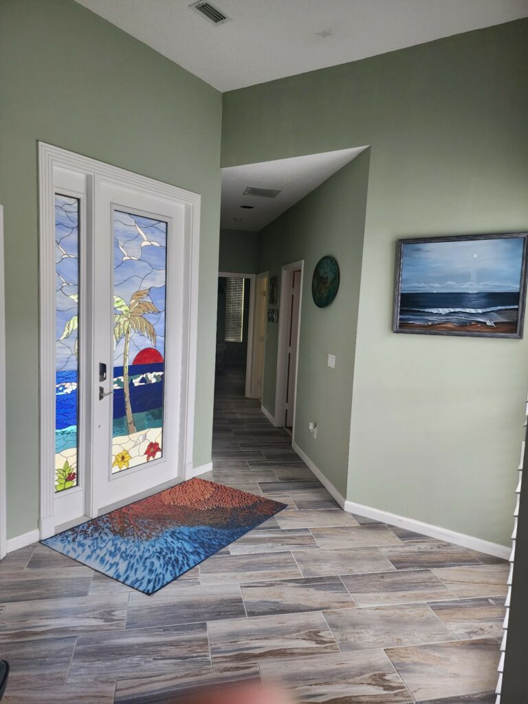 A set of white-framed double entry doors, each featuring a stained glass panel. The combined design depicts a vibrant tropical beach scene with a central palm tree, a large red sun setting over blue ocean waves, sandy shores with seashells and red and yellow hibiscus flowers, and white seagulls flying in a light blue sky.