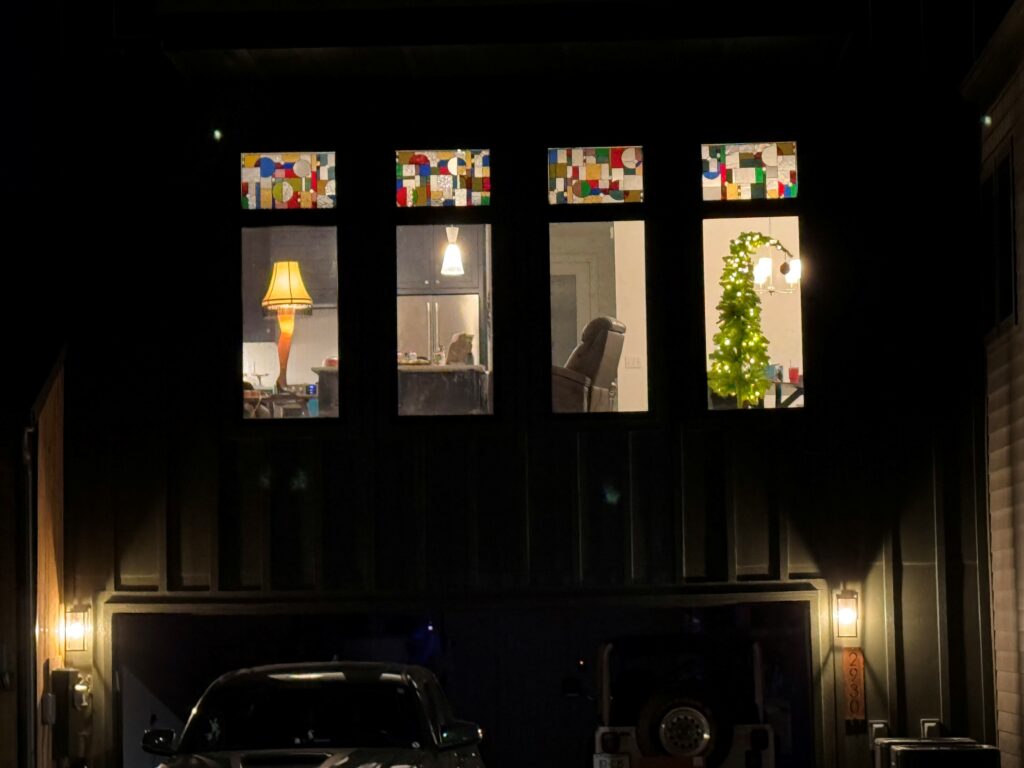 Four rectangular stained glass transom panels, each featuring an abstract geometric mosaic design with colorful squares, rectangles, and circular elements in red, blue, green, yellow, and clear glass, illuminated at night above home windows.