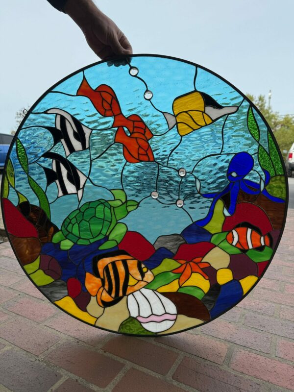 A vibrant, round stained glass panel depicting an underwater scene with various marine life. A green sea turtle, blue octopus, clownfish, striped angelfish, and other colorful fish swim among colorful rocks, a white shell, and green kelp, all set against textured blue water with clear glass bubbles.