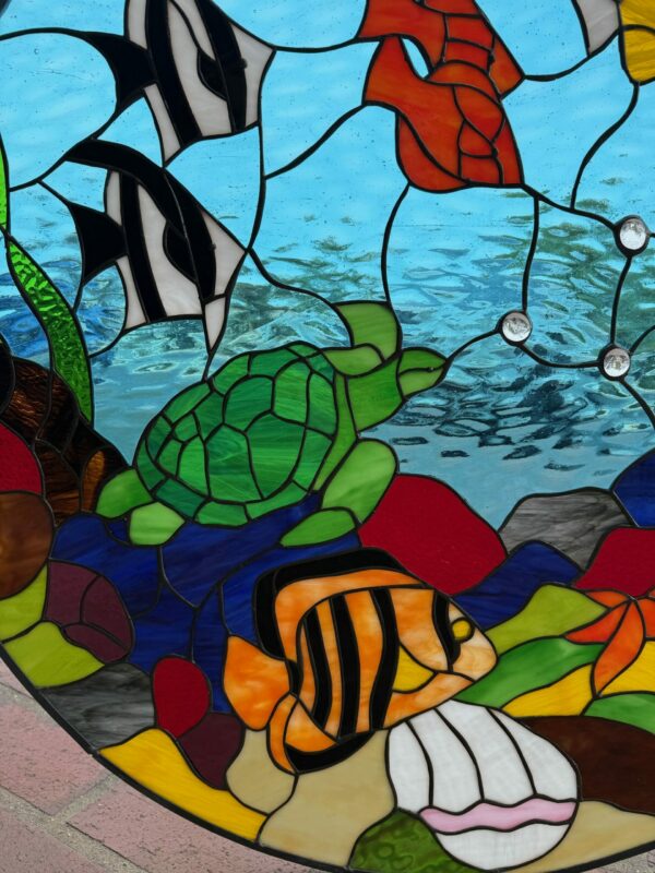 A vibrant stained glass panel depicting an underwater coral reef scene with a green sea turtle, two striped tropical fish, red and green coral, and clear round jewels in blue water.