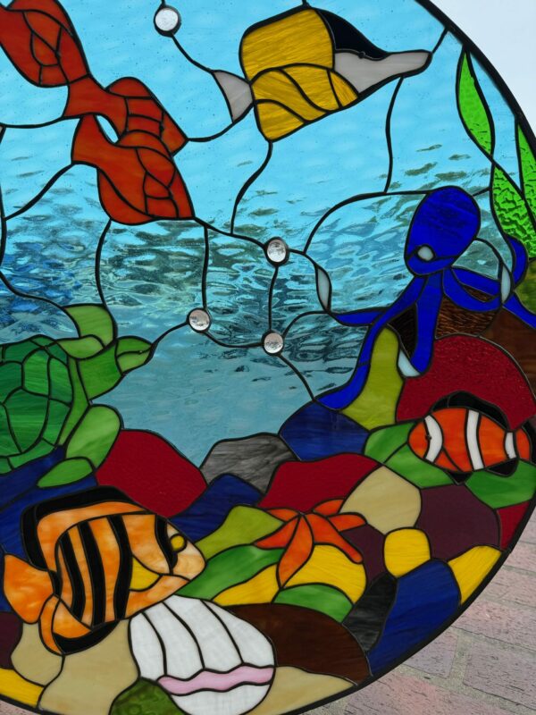 A circular stained glass panel depicting an underwater coral reef scene with colorful tropical fish, a green sea turtle, and a blue octopus.