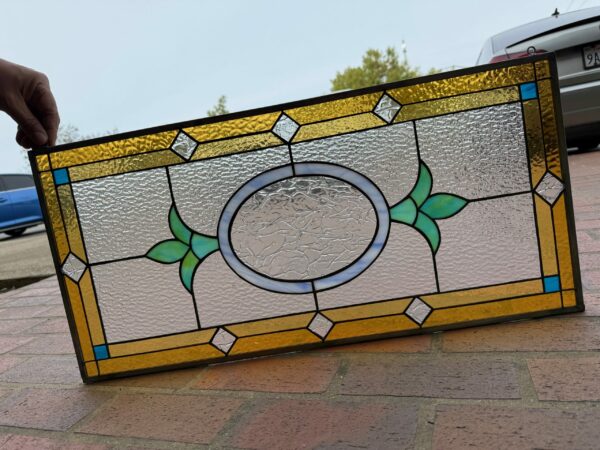A rectangular leaded stained glass panel with a central textured clear oval, flanked by green leaf designs, surrounded by a border of textured amber glass, with small clear diamond shapes and blue corner squares.