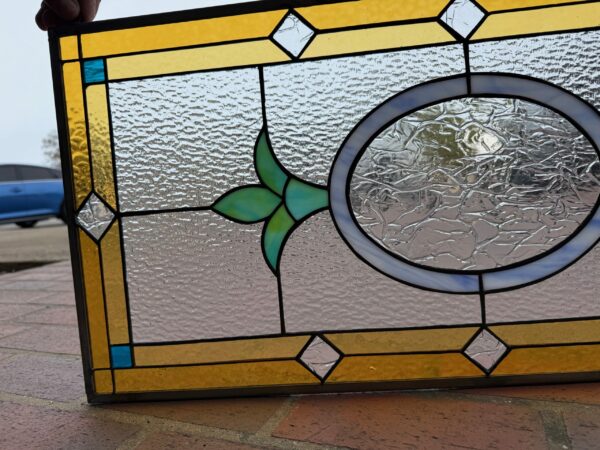 A rectangular leaded stained glass panel features a wide amber-yellow border, framing a field of clear, heavily rippled privacy glass. A central clear circle, bordered with opalescent light blue glass, is accented by a stylized four-petal floral design in shades of green and teal. Small clear textured diamond inserts are positioned along the amber border, and the glass within the central circle has a crinkled texture.