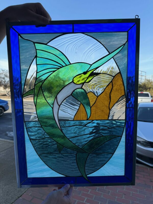 A rectangular stained glass panel featuring a green and blue marlin leaping from textured blue water, framed by a deep royal blue border. The background shows golden islands and a clear wavy sky.