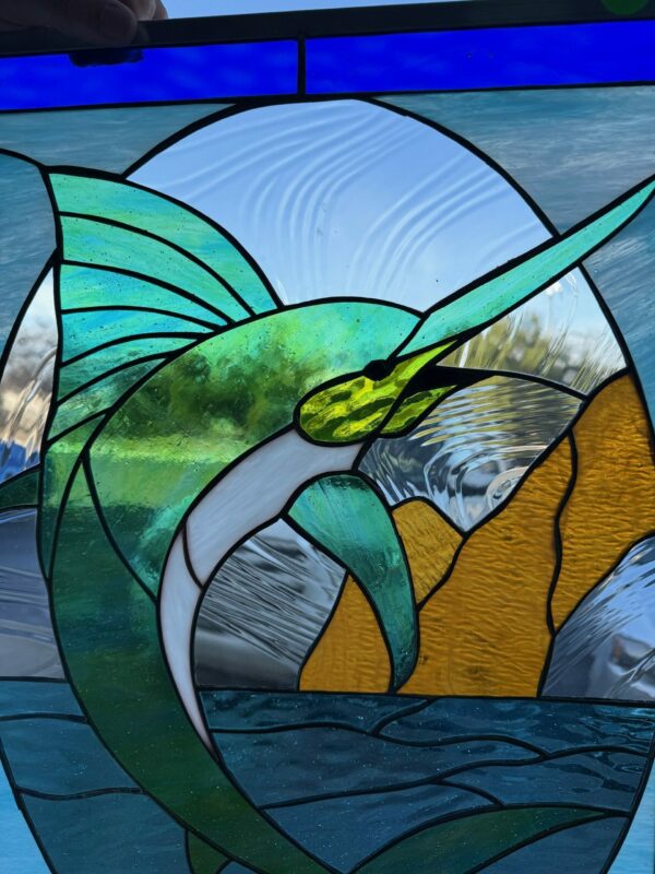 A vibrant, leaded stained glass panel depicting a green and blue sailfish leaping out of blue water, with a textured clear sky and amber textured rocks in the background.