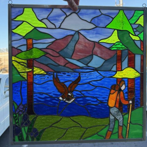 A rectangular stained glass panel depicting a vibrant mountain and lake landscape. The scene features blue and white sky, green pine trees, distant reddish-brown and blue mountains, a textured dark blue lake, green ground with purple iris flowers, a brown eagle flying over the lake, and an orange-clad hiker with a backpack and walking stick on the right. The panel is held by a hand against a clear blue sky.