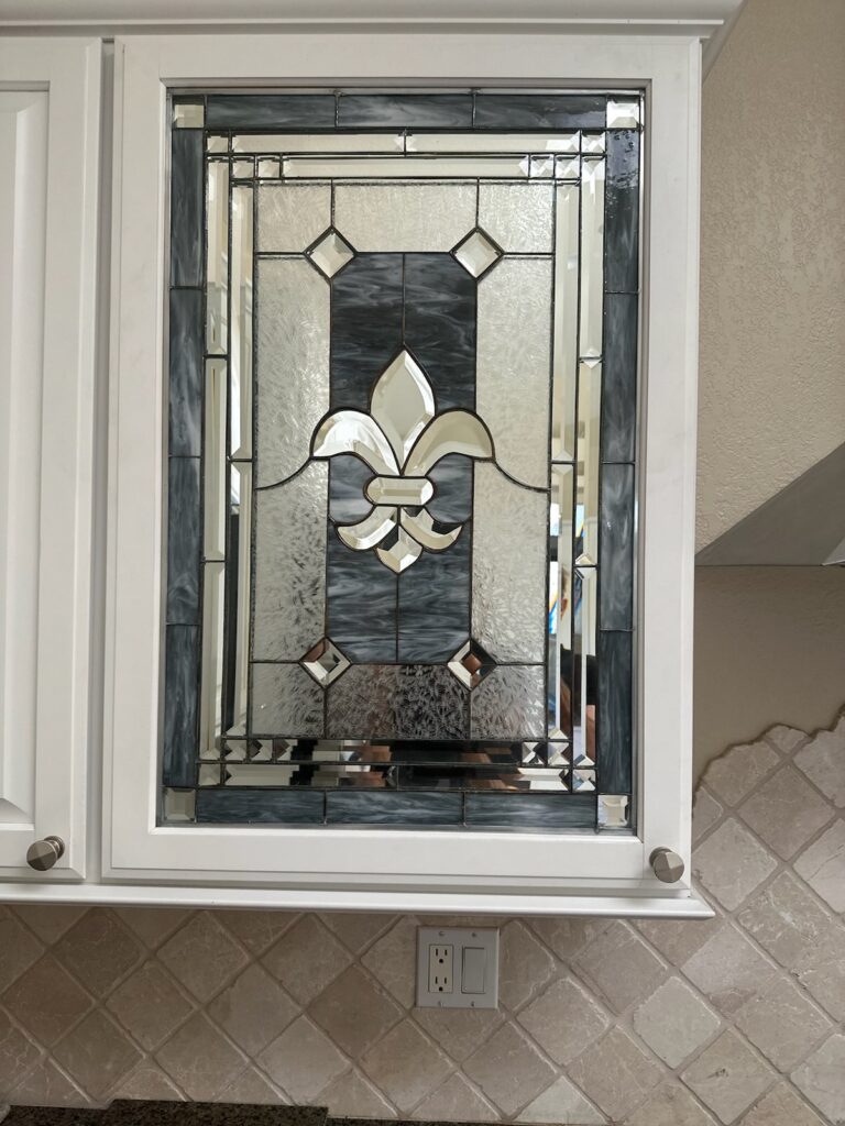 A rectangular stained glass panel installed in a white kitchen cabinet door, featuring a central clear beveled fleur-de-lis, bordered by clear textured glass and dark gray mottled art glass.