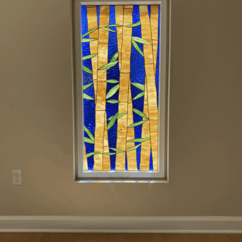 A rectangular stained glass window featuring golden-yellow bamboo stalks with green leaves, set against a vibrant blue background, installed in a neutral wall with a white frame.