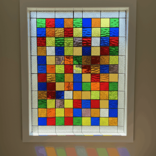 A rectangular, insulated vinyl window with a colorful stained glass panel inside. The design is a grid of multi-colored squares in shades of red, blue, green, and yellow, surrounded by a clear, textured border.