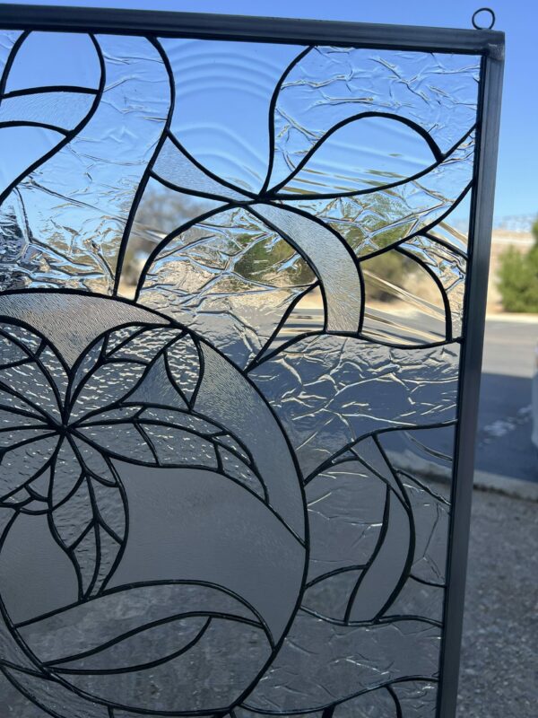 A rectangular clear stained glass panel featuring an abstract design with a central floral motif and flowing organic patterns, framed in dark metal.