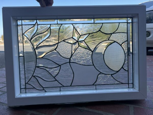 A rectangular, white-framed, insulated stained glass window featuring a contemporary clear design with abstract sun, star, and crescent moon shapes, utilizing various textured and beveled glass.