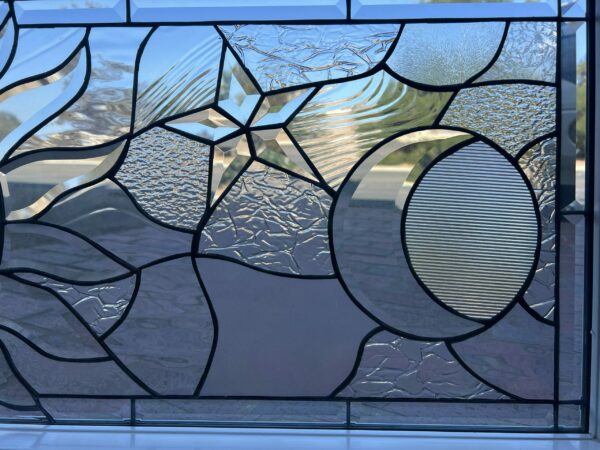 A clear leaded stained glass panel featuring a central beveled star, a textured crescent moon, and a fluted circular element, surrounded by various clear textured glass patterns like crinkled and rippled glass.