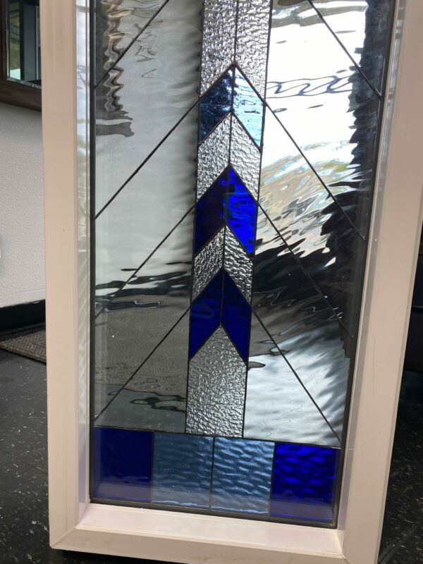A rectangular stained glass panel in a white frame, featuring clear rippled glass with a central vertical stripe of alternating deep blue and silver textured chevron patterns, and blue rectangular sections at the bottom.