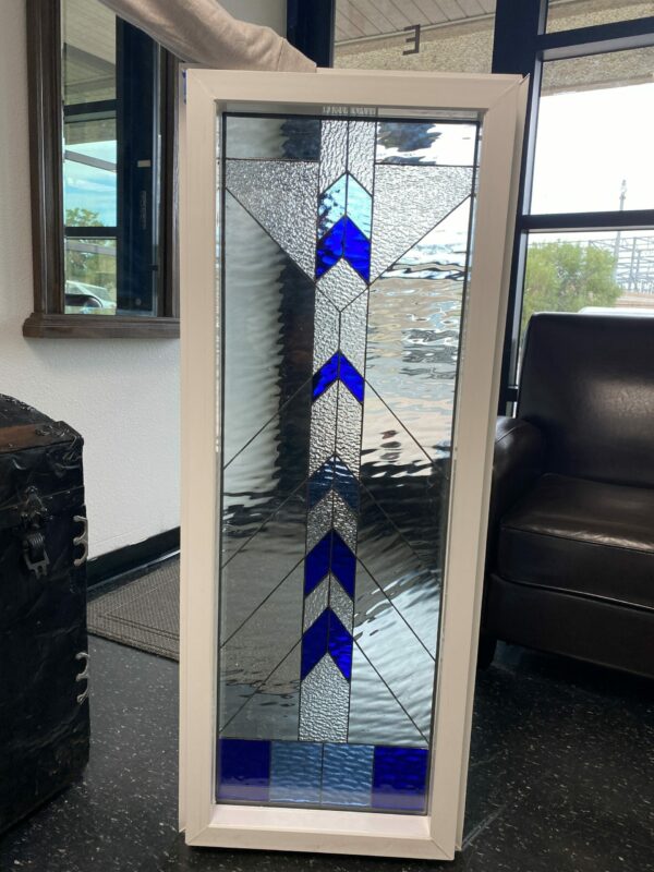 A tall, rectangular stained glass panel in a white frame, featuring a bold geometric design with black lead lines. The panel primarily uses various clear textured glasses, including water glass, hammered, and glue chip, which diffuse light. Vibrant deep blue and light blue glass pieces are arranged in a repeating pattern of upward-pointing arrows or chevrons that run vertically through the center of the panel.