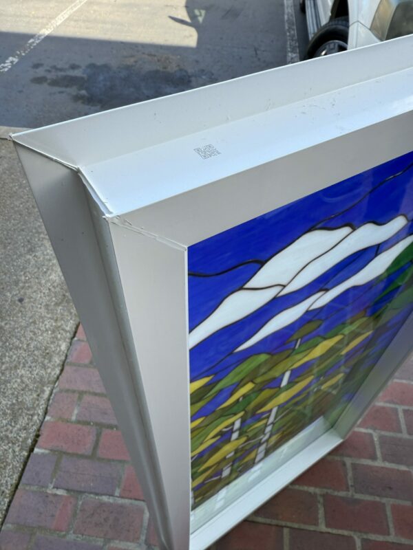 A rectangular stained glass panel set within a white, robust insulated frame. The vibrant design features a blue sky, flowing white clouds, and undulating green and yellow abstract foliage. The panel is seen from an elevated angle, resting on red brick pavers with an asphalt surface in the background. A QR code is visible on the upper frame.