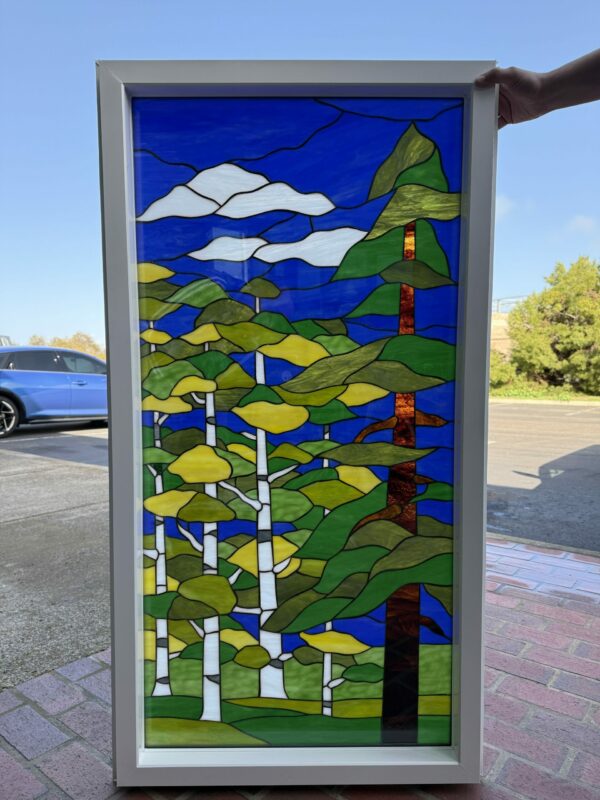 A rectangular stained glass panel depicting a stylized woodland scene with white birch trees, green and yellow foliage, a dark pine tree, a bright blue sky, and white clouds, framed in light gray.