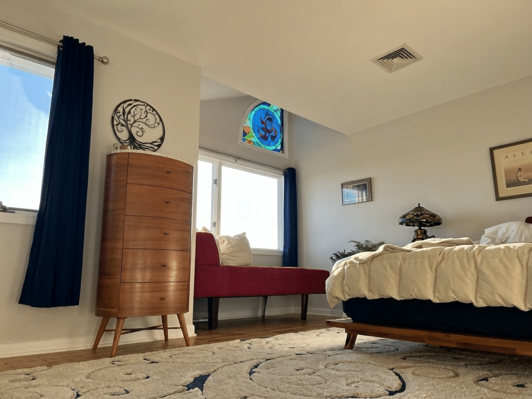 An arched stained glass transom window featuring a vibrant abstract circular design in shades of blue, red, green, and orange, positioned above a larger rectangular window in a brightly lit room.