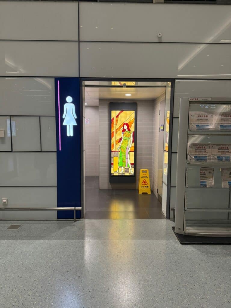 A tall, rectangular stained glass panel depicting a woman with red hair wearing a green cheongsam and holding a yellow suitcase, set against an abstract background of warm yellow, orange, and red glass.