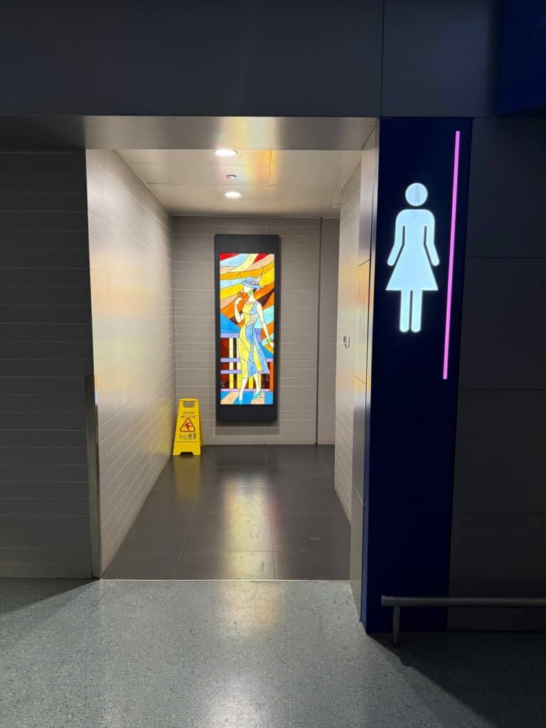 A tall, rectangular stained glass panel illustrating a stylized woman in a wide-brimmed hat and flowing dress, crafted with vibrant geometric pieces in red, orange, yellow, blue, and green.