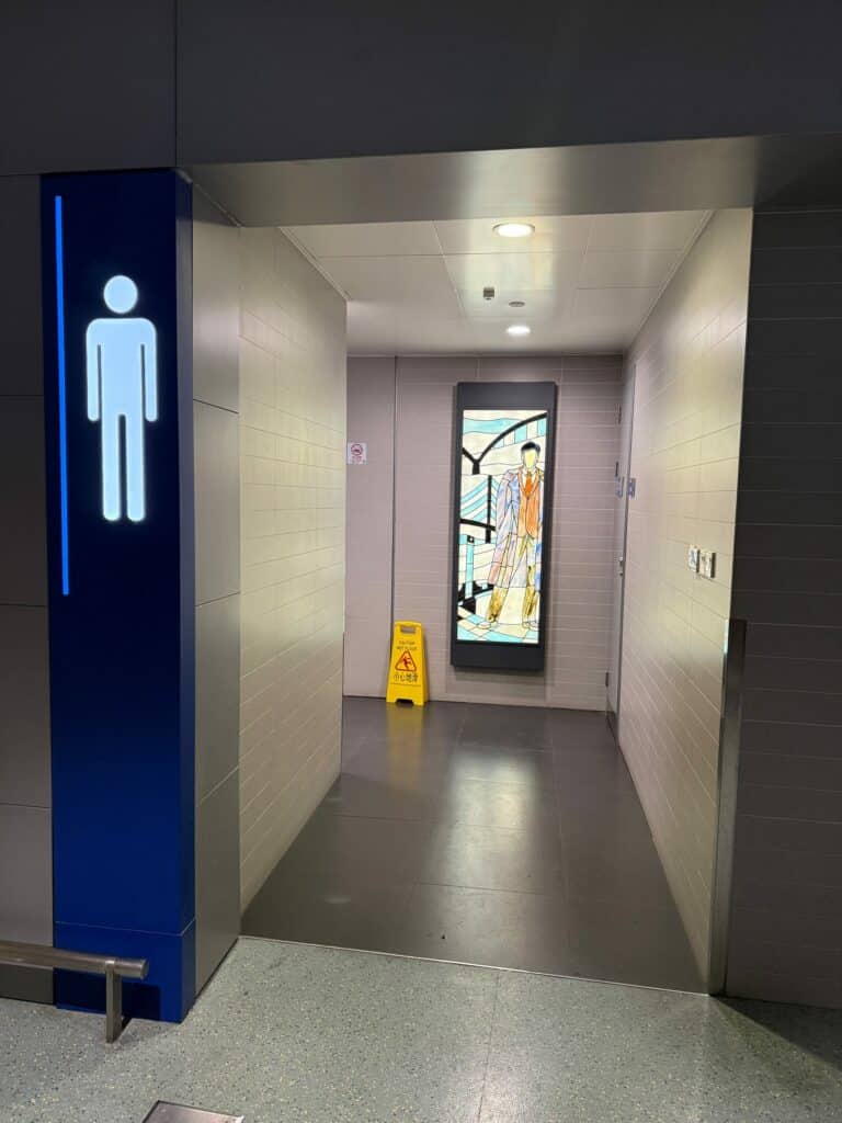 A tall, vertical stained glass panel depicting a stylized man in a suit with an orange tie, set against a geometric blue background, framed in black, and installed in a modern tiled hallway.