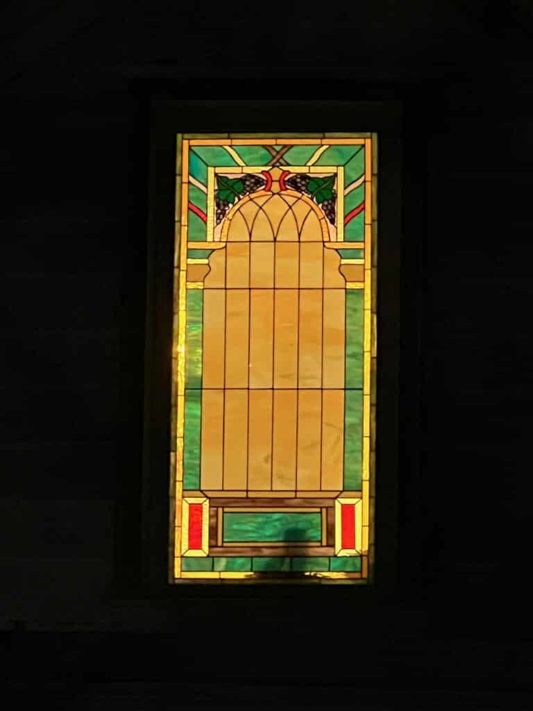A tall, rectangular stained glass panel with a central amber arched motif, green and yellow geometric borders, and a top design of purple grapes and green leaves.