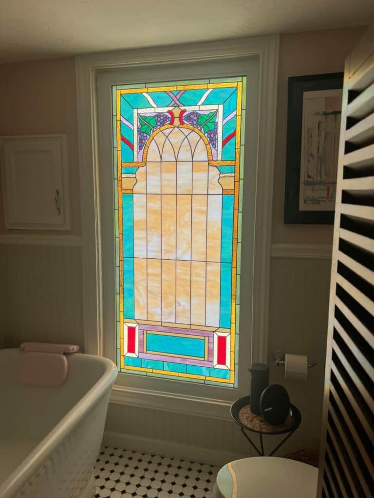 A rectangular stained glass window with an arched top section featuring purple grape clusters, green leaves, and clear amber glass, flanked by turquoise and red accents, installed in a bathroom.