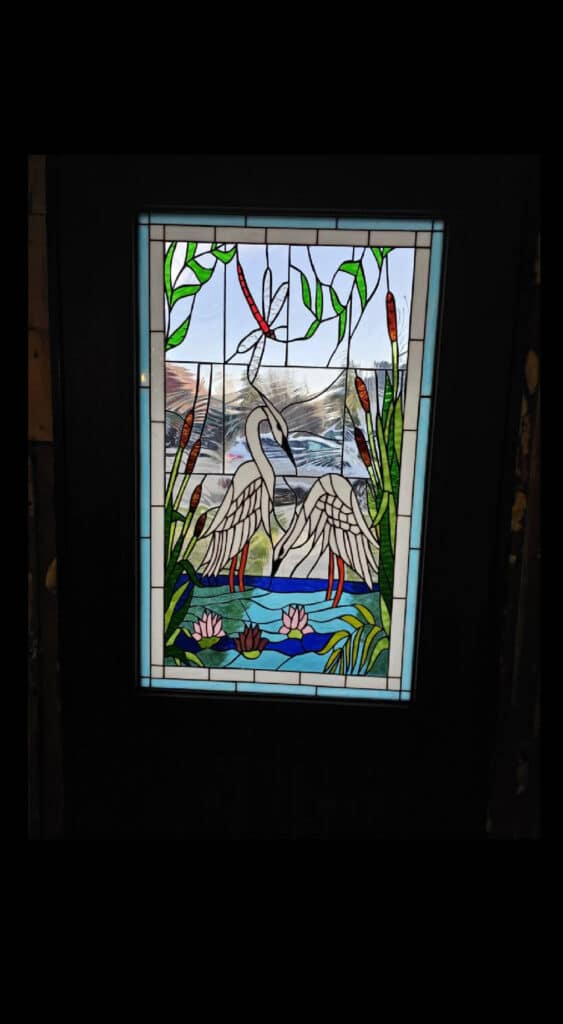 Rectangular stained glass panel depicting two white egrets wading in a blue pond with pink water lilies, green cattails, and a red dragonfly overhead.