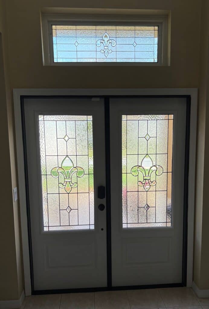 A set of white double entry doors with matching rectangular transom window above. Each features a clear textured leaded stained glass panel with a geometric pattern, diamond accents, and a central fleur-de-lis design in light green and amber hues.