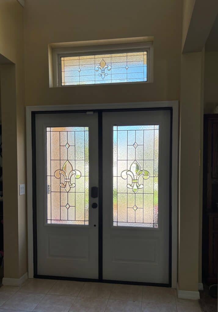 A pair of white double entry doors with leaded stained glass panels featuring a central Fleur-de-lis design, clear textured glass, and small clear bevels, topped by a matching rectangular stained glass transom window.