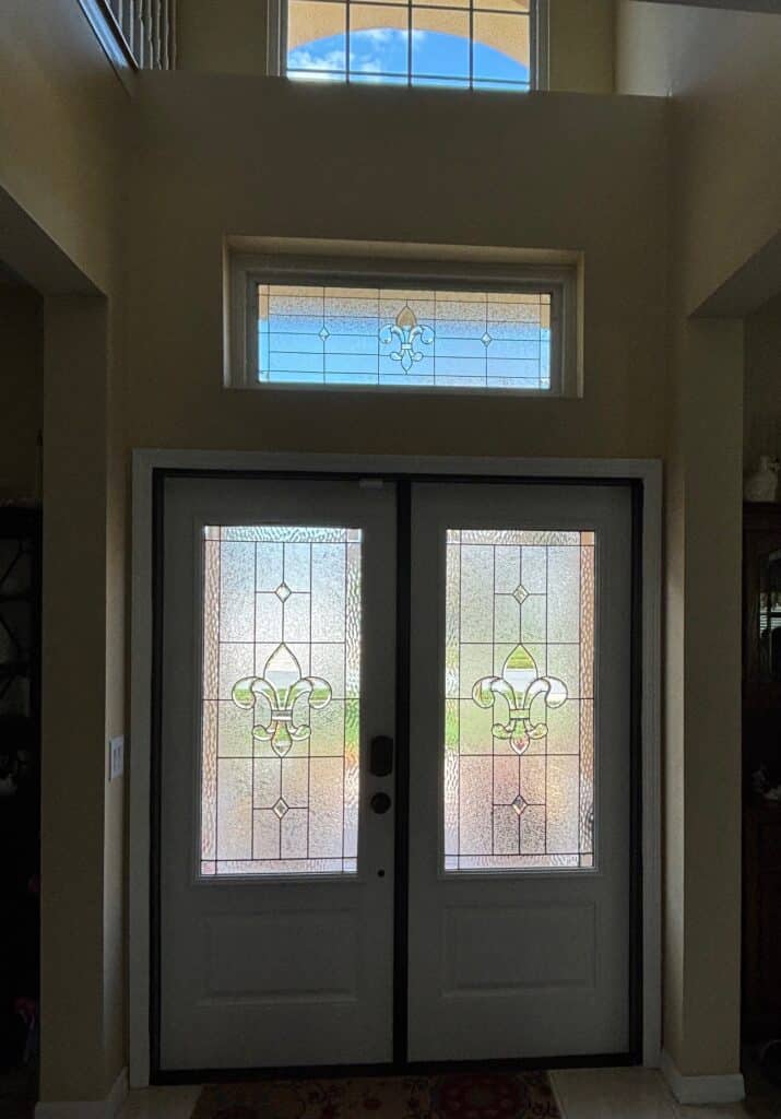 A home entryway featuring white double doors with leaded clear textured stained glass inserts and a matching rectangular transom window above, all showcasing a central Fleur-de-lis design.
