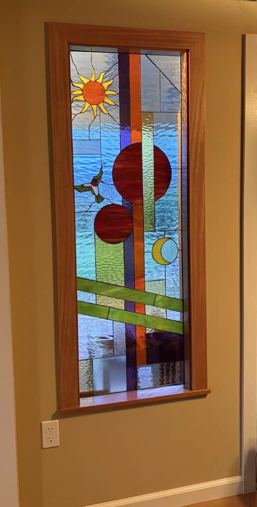 A tall rectangular stained glass panel in a wooden frame, featuring a yellow sun, a red and green hummingbird, three red circles, a yellow crescent moon, and abstract geometric patterns in blue, green, orange, purple, and textured clear glass.