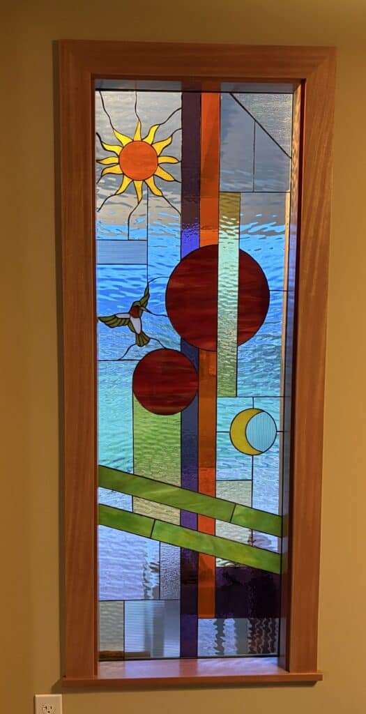 A vertical rectangular stained glass panel in a light wooden frame, depicting an abstract scene with a sun, hummingbird, planets, and moon. The top left features a yellow sun with an orange center. Below it, a green and red hummingbird is in flight. On the right, there are two large reddish-brown circular elements and a smaller yellow crescent moon with blue horizontal lines. The background consists of various textured blue, green, orange, and purple rectangular and diagonal glass sections, many with a wavy water-like texture.