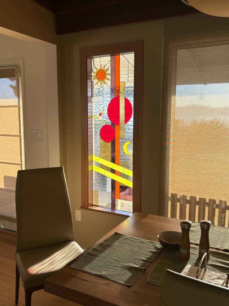 A rectangular stained glass window with a modern abstract design featuring a yellow sun, two large red circles, a crescent moon, a white bird, and varied clear textured glass, set in a wooden frame.