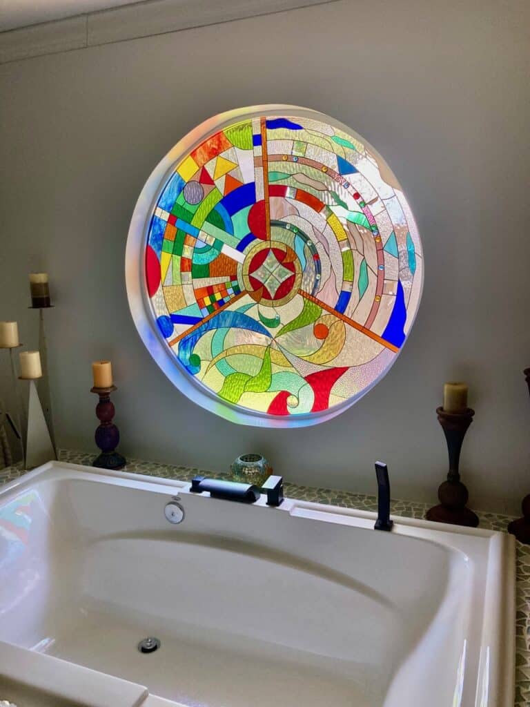 A large, round stained glass window with a vibrant, complex abstract geometric design in multiple colors, featuring a central square motif, installed above a white built-in bathtub.