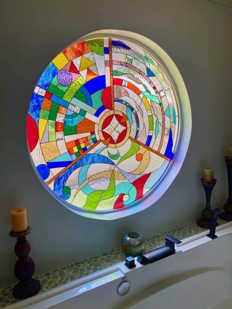 A large, circular stained glass window in a white frame, displaying a vibrant, abstract geometric pattern with a central starburst and radiating colorful segments, installed above a bathtub.