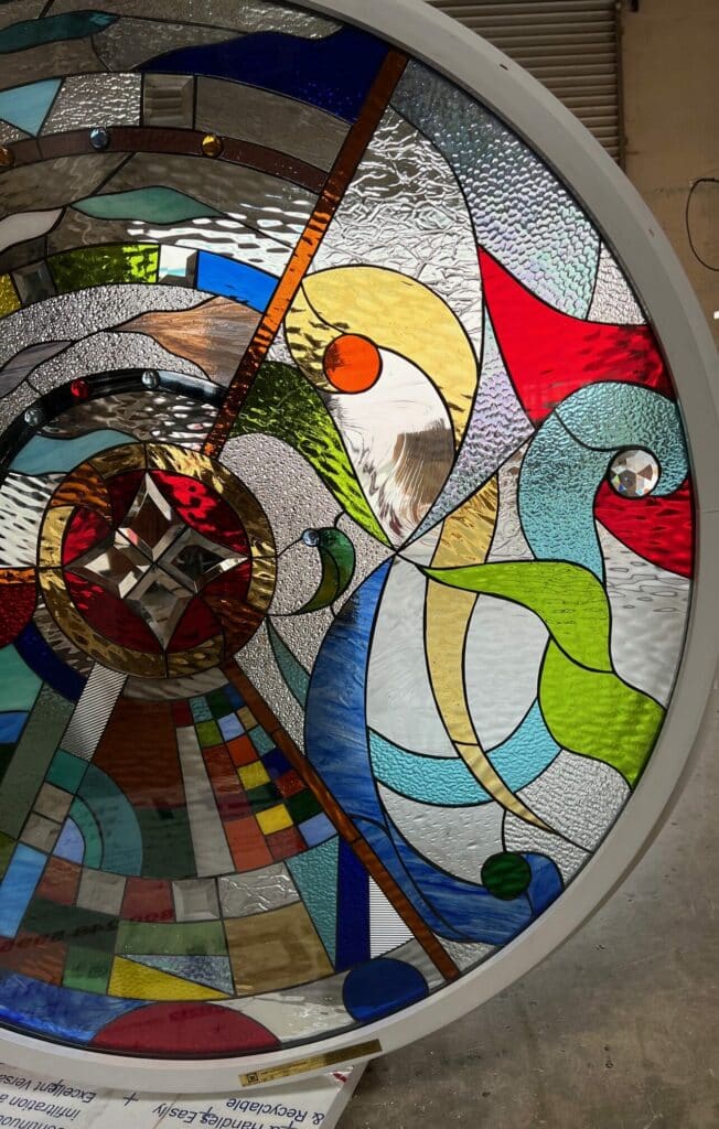 A large, round stained glass panel featuring a complex, abstract and geometric design in a wide spectrum of colors including reds, blues, greens, yellows, and clear textured glass, with prominent faceted jewels and beveled sections, set in a white circular frame.