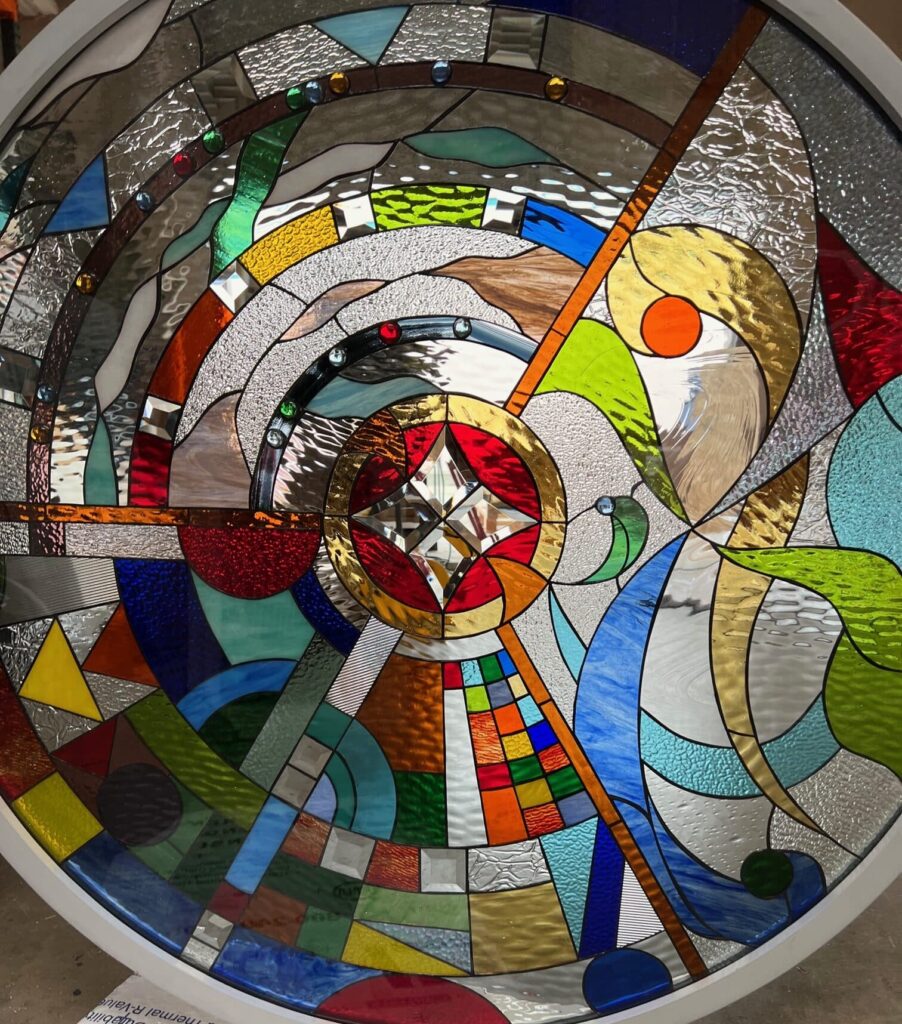 A circular stained glass panel with a vibrant abstract, multi-colored design, featuring a central clear bevel cluster, varied textured clear glass, and embedded glass jewels, all within a white frame.