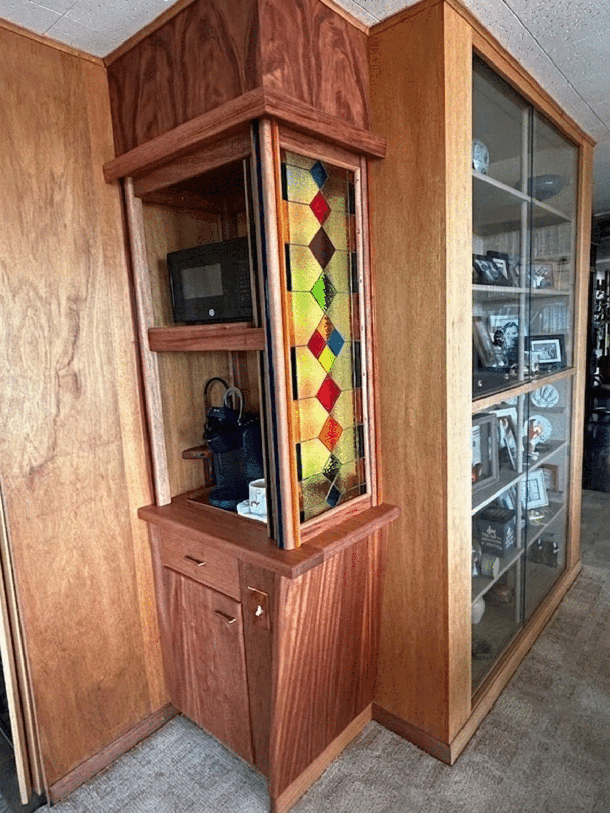 A tall, rectangular leaded stained glass panel featuring a repeating geometric diamond pattern in multiple colors including amber, green, red, and blue, set within a custom wooden cabinet.