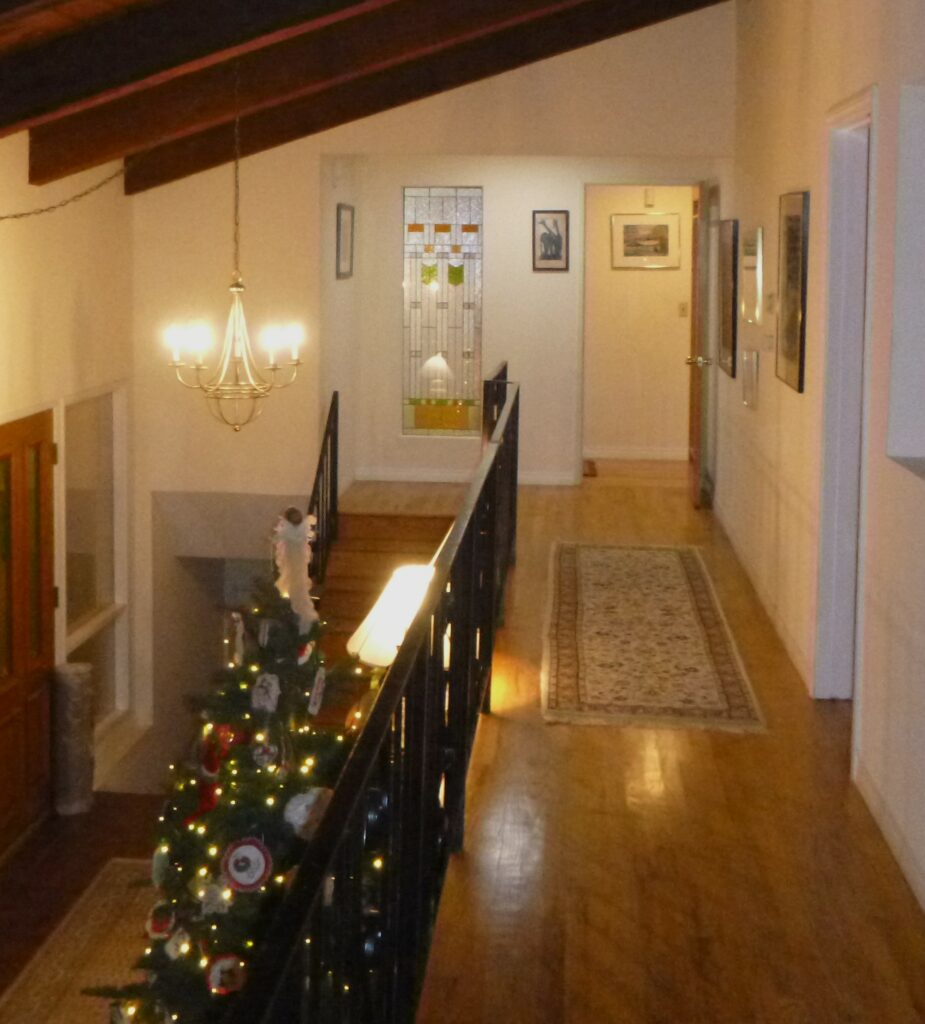 A tall rectangular leaded stained glass panel with clear textured glass, amber horizontal accents, and two green inverted V-shaped elements, installed in a white wall at the end of a hallway.