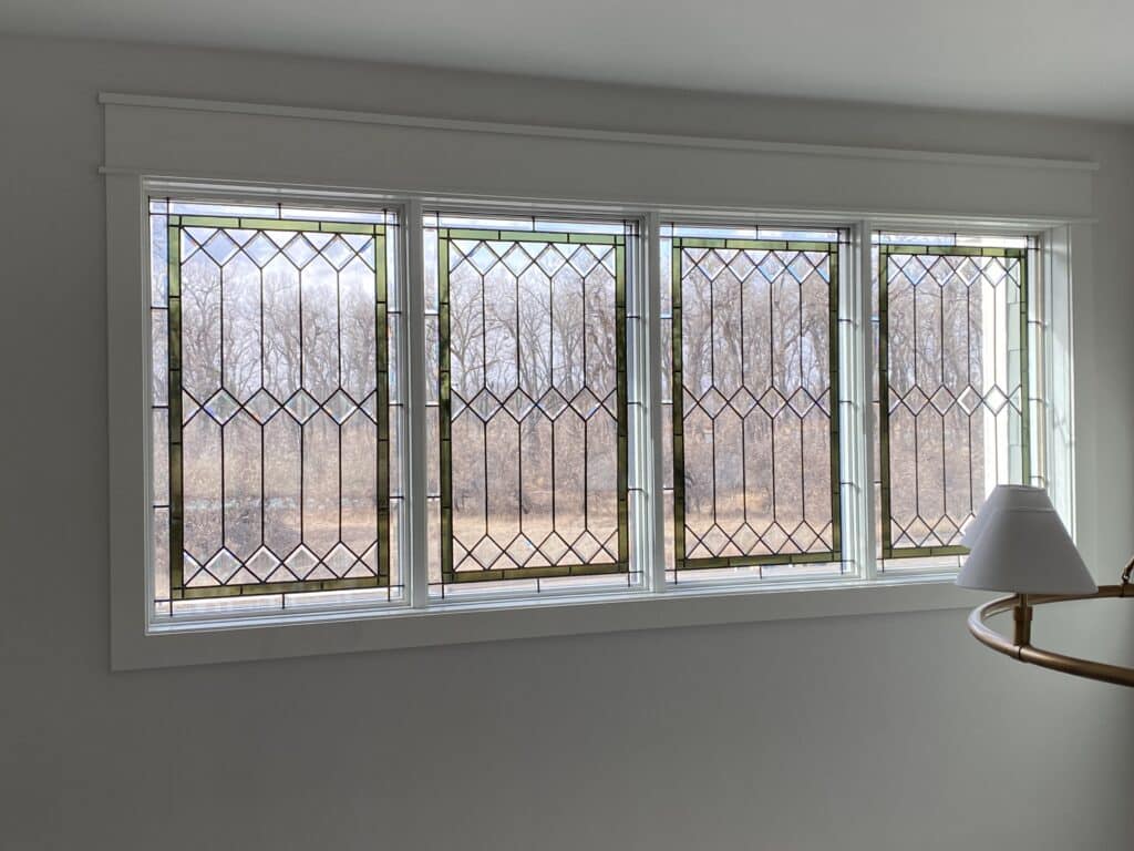 A long, rectangular transom window composed of four leaded stained glass panels, each featuring a geometric design with clear diamond bevels and light green border accents, set in a white frame, with a view of bare trees outside.