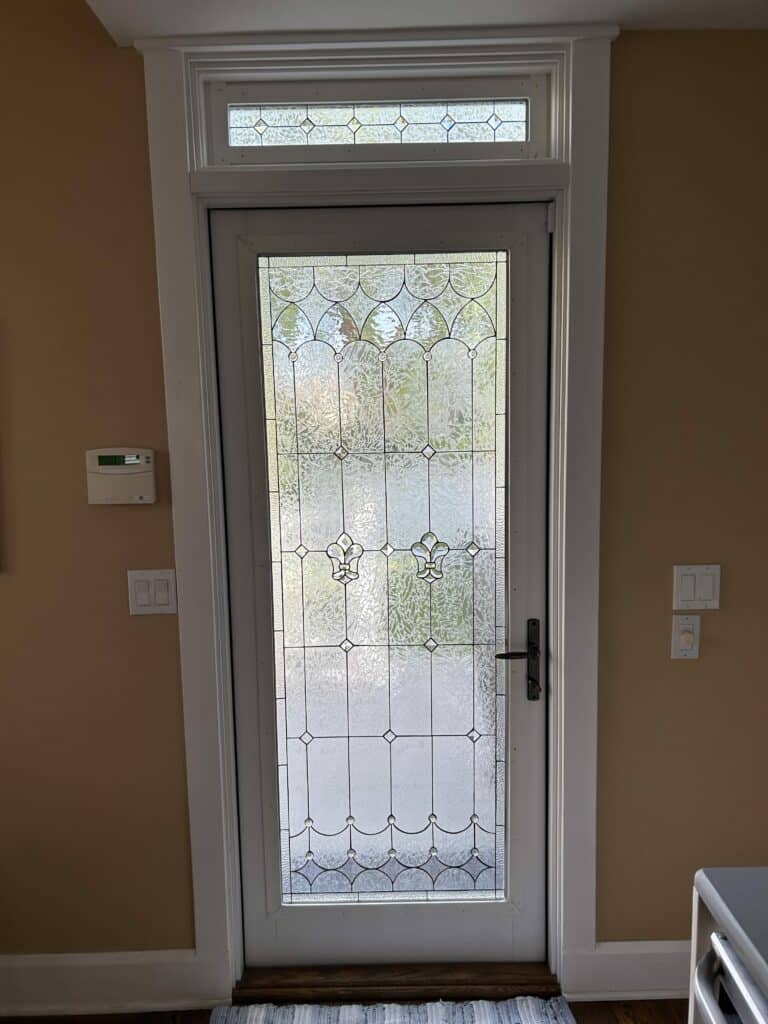 A white full-lite entry door and matching rectangular transom window, both featuring clear textured privacy glass with a leaded geometric pattern. The door panel includes two central fleur-de-lis motifs and small diamond accents, while the transom displays a complementary grid design with small beveled squares.