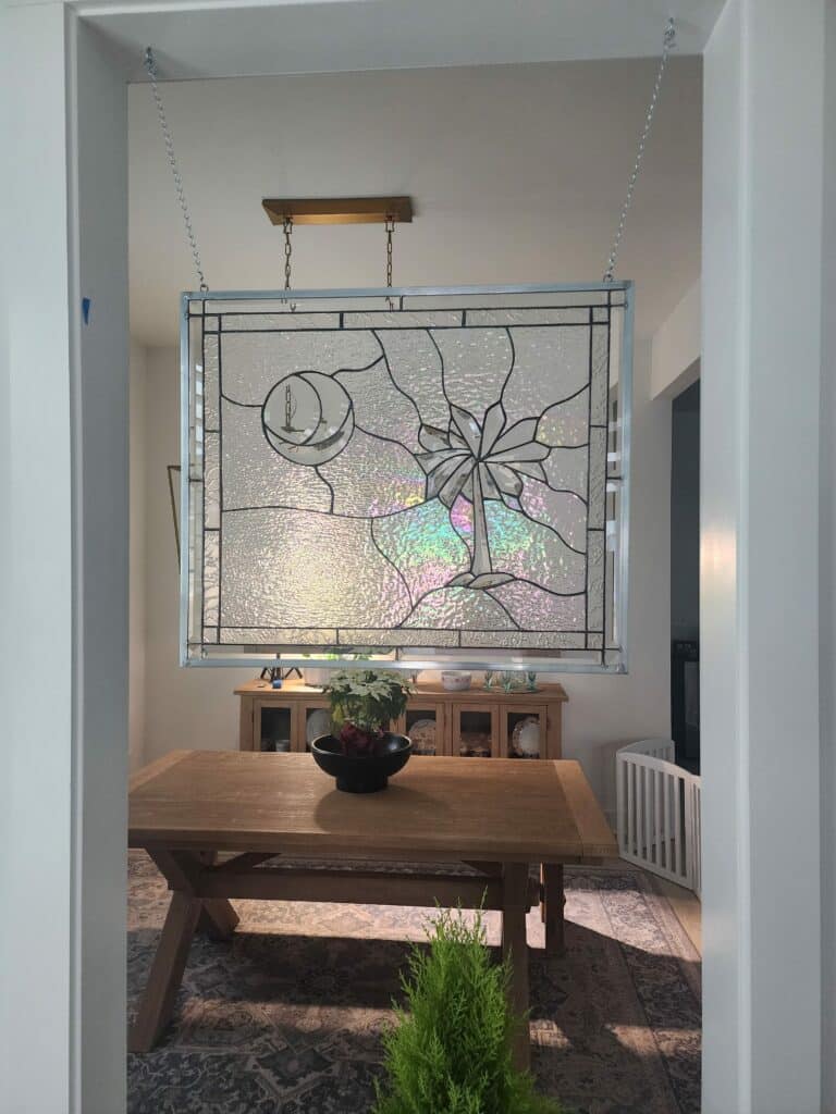 A rectangular, clear textured stained glass panel depicting a Palmetto tree and a crescent moon with a lighthouse, suspended by chains in a doorway. The panel incorporates clear beveled glass for the central elements and a mix of textured clear and iridescent glass, framed in silver-toned metal came.