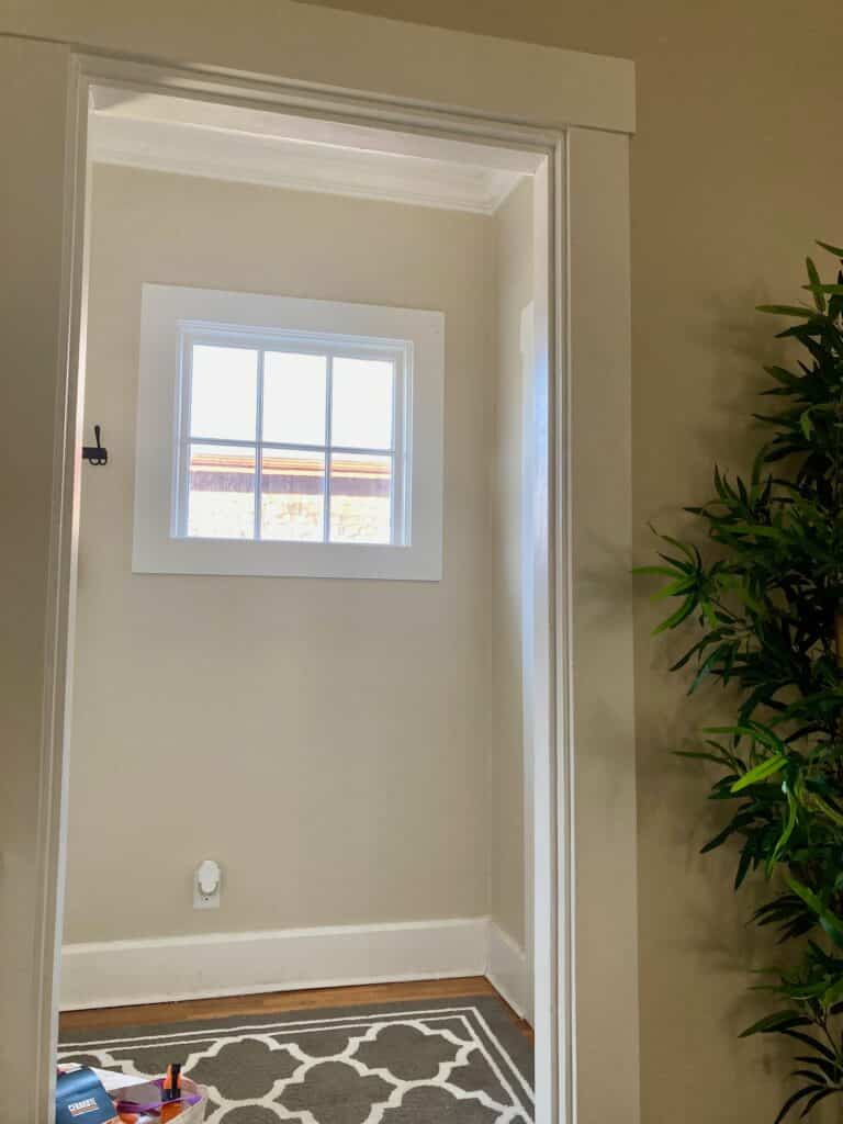 A small, white-framed, multi-pane window in a bright, neutral-colored hallway, ideal for a custom stained glass privacy panel, with a decorative rug and a tall green plant.