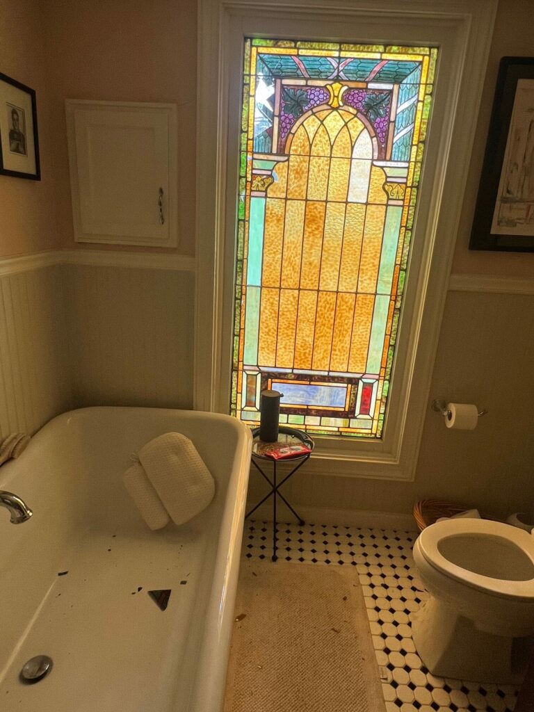 A tall, rectangular leaded stained glass window with an arched top, featuring purple grapes, green leaves, and amber textured glass. The lower section includes blue and red glass accents.