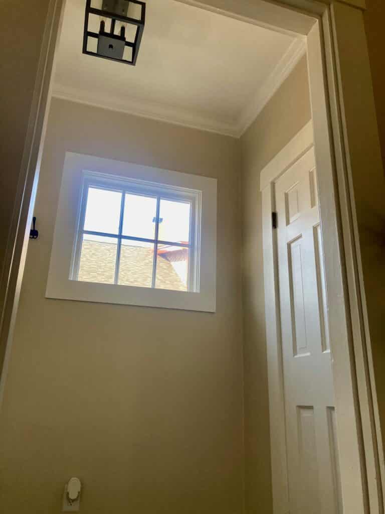 A plain clear grid window in a neutral-painted hallway, viewed from a low angle. A dark modern light fixture is visible on the ceiling, and a white paneled door is partially open on the right.