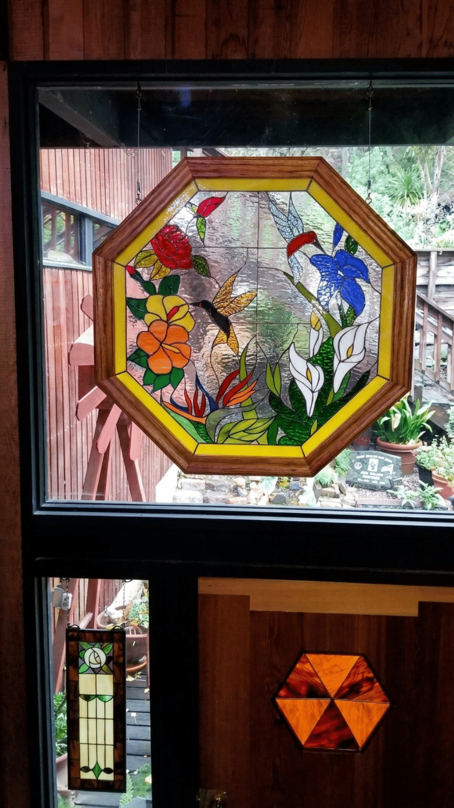 An octagonal, wood-framed stained glass panel featuring two hummingbirds surrounded by a vibrant arrangement of red roses, hibiscus, blue flowers, and white calla lilies.