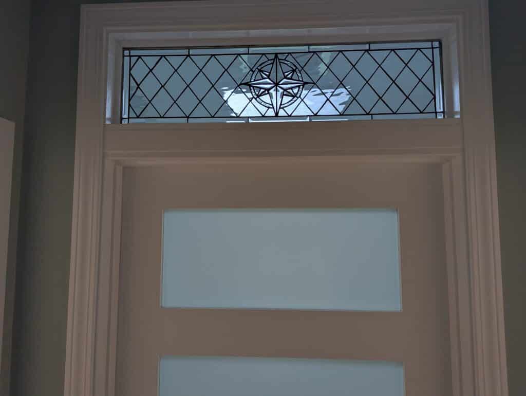 A rectangular clear beveled and leaded glass transom window featuring a central compass rose medallion. The background is composed of diamond-shaped leaded glass, framed by a plain white door frame above a door with two frosted glass panels.