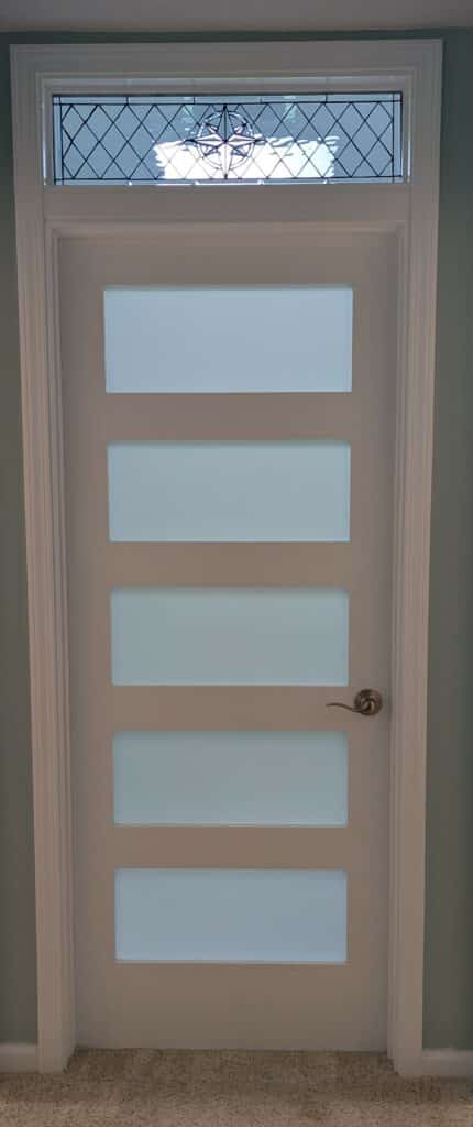 A white interior door with five horizontal rectangular frosted glass panels below a rectangular leaded clear glass transom. The transom features a diamond grid pattern and a central compass rose medallion.